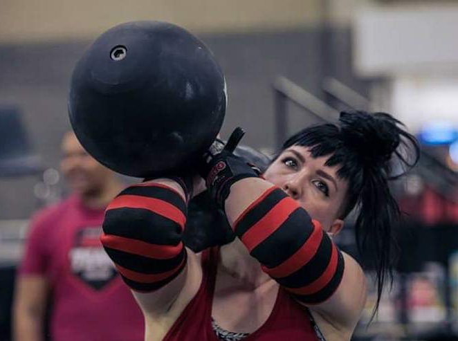 The author holds a giant lifting dumbbell to one shoulder, preparing to press it overhead with one arm.